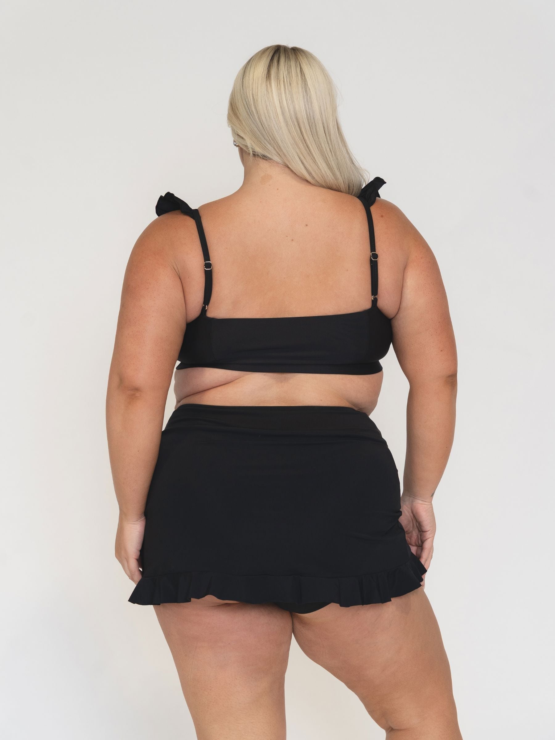 view from behind of a woman wearing a black ruffle bikini and ruffle skirt on a white background