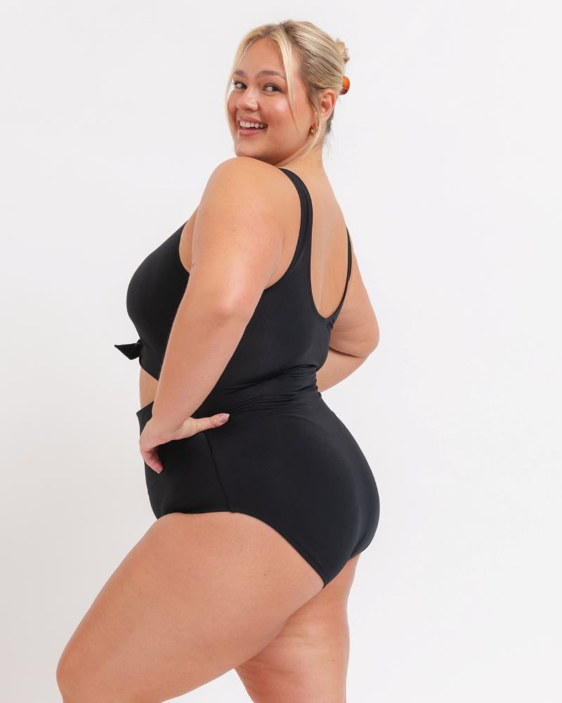side view of a woman wearing a black knotted one piece with a white background