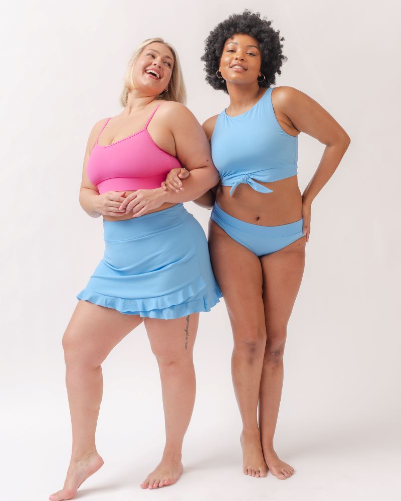 Photo of two women one wearing a pink cropped swim top with a light periwinkle swim skirt the other wearing a light periwinkle cropped swim top with a light periwinkle classic swim bottom