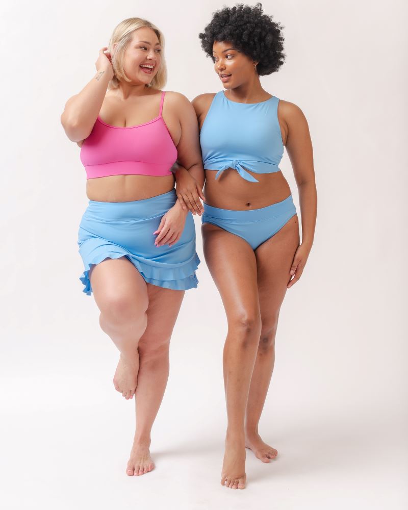 Photo of two women one wearing a pink cropped swim top with a light periwinkle swim skirt the other wearing a light periwinkle cropped swim top with a light periwinkle classic swim bottom