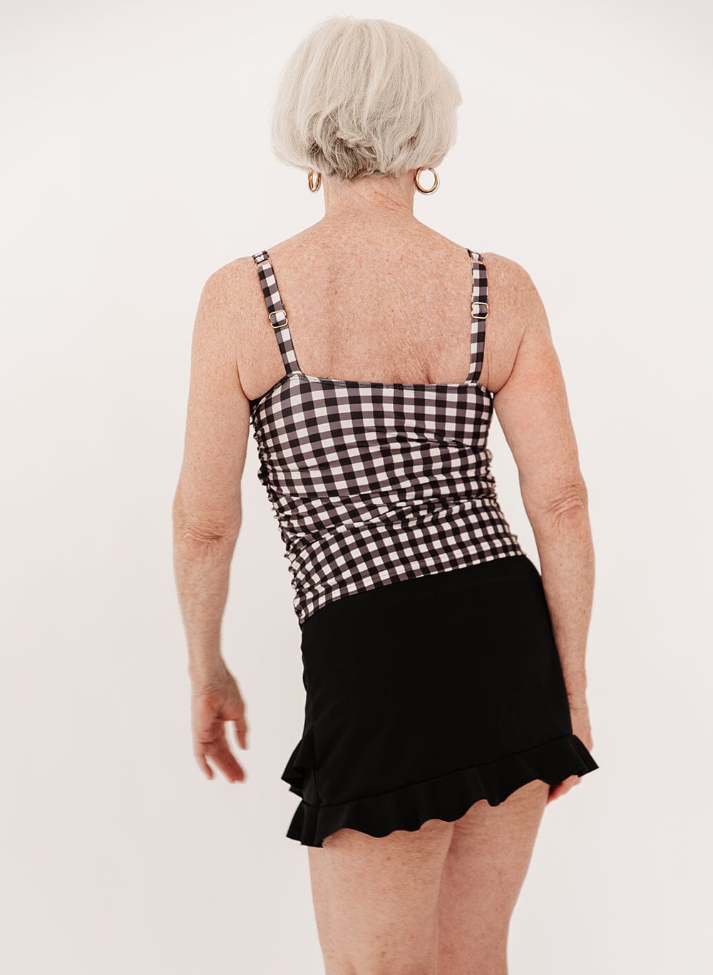  woman wearing a black high-waist ruffle swim skirt with a black gingham square neck tankini swim top side view